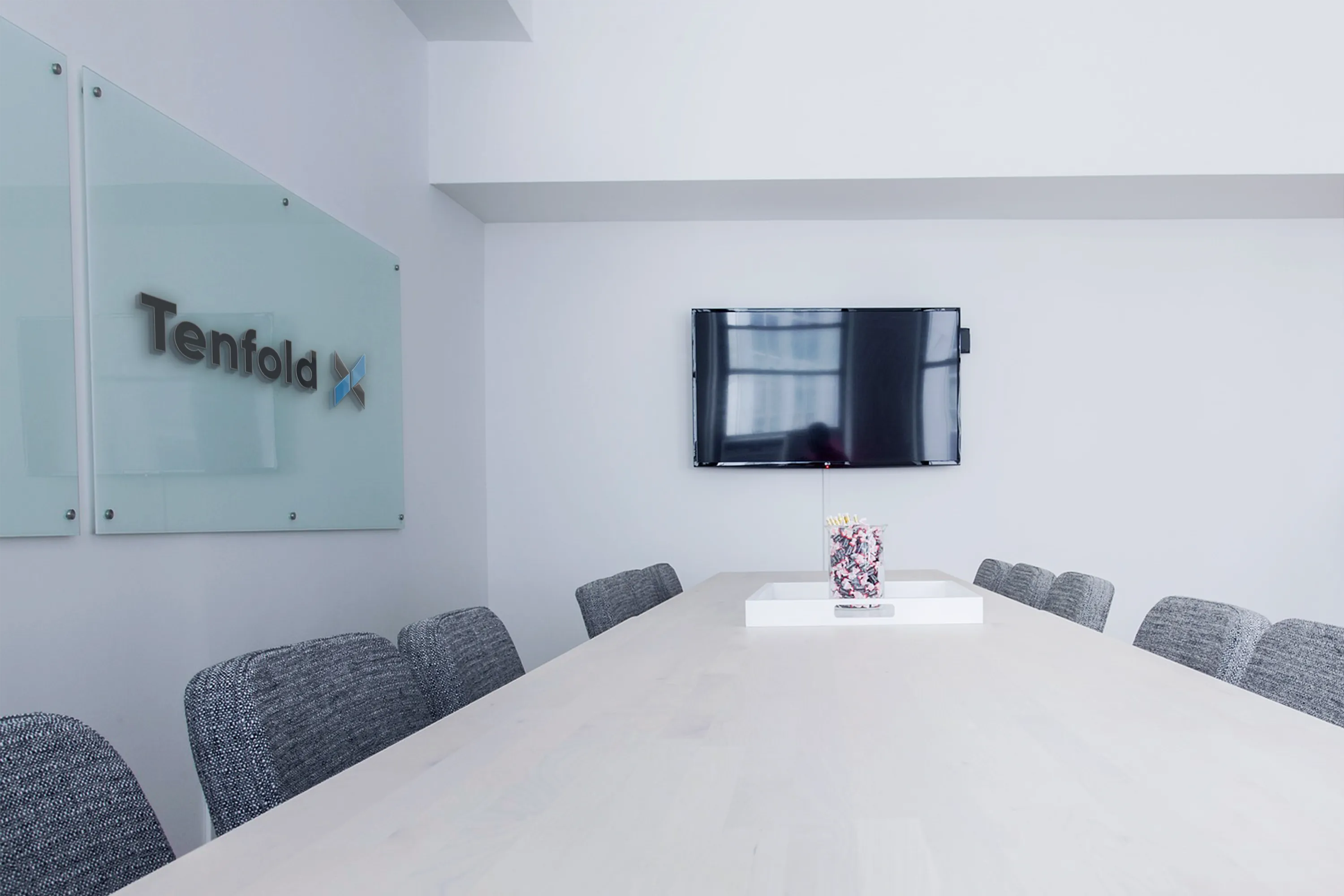 Modern conference room with a long table, gray chairs, a wall-mounted TV, and a glass sign reading 'Tenfold' on the wall.