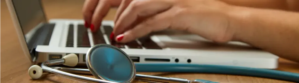 Hands typing on laptop with stethoscope on wooden desk, suggesting healthcare or medical professional at work